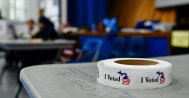 A roll of "I Voted" stickers lays on a grey table, as voting activity takes place out of focus in the background.