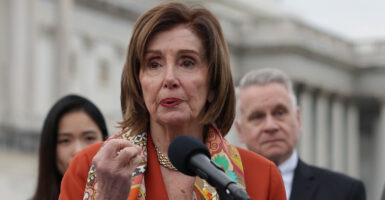 Nancy Pelosi stands at a microphone wearing and orange blazer.