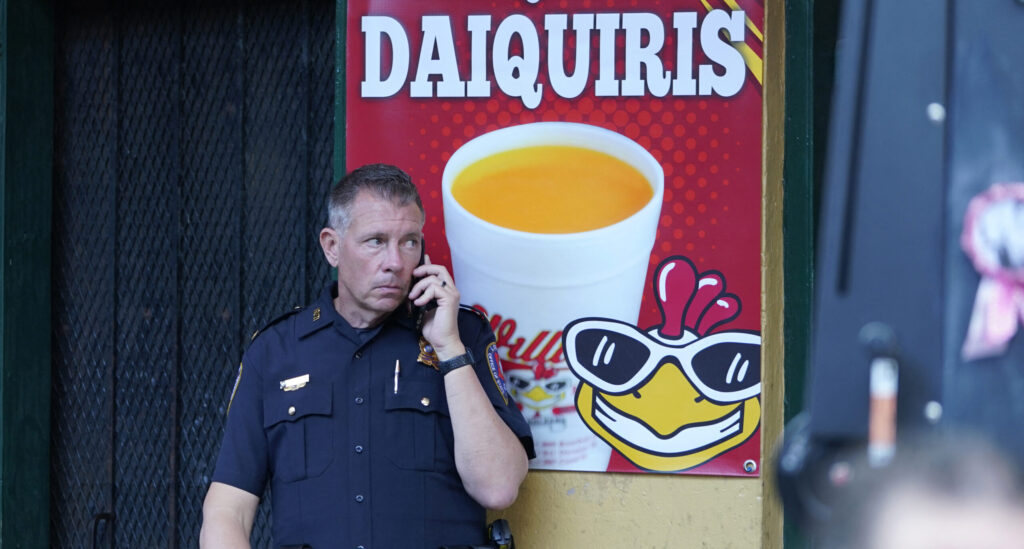 A New Orleans police officer in the French Quarter stands in front of a sign for daiquiris, including a logo of a smiling chicken head in sunglasses.