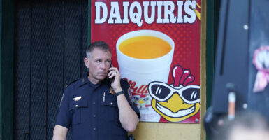 A New Orleans police officer in the French Quarter stands in front of a sign for daiquiris, including a logo of a smiling chicken head in sunglasses.
