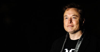 Elon Musk stands in a dark room wearing black clothing.
