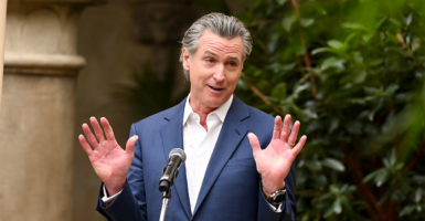 California Gov. Gavin Newsom in a blue suit jacket and no tie standing in front of a microphone outdoors with his palms raised like he's surrendering