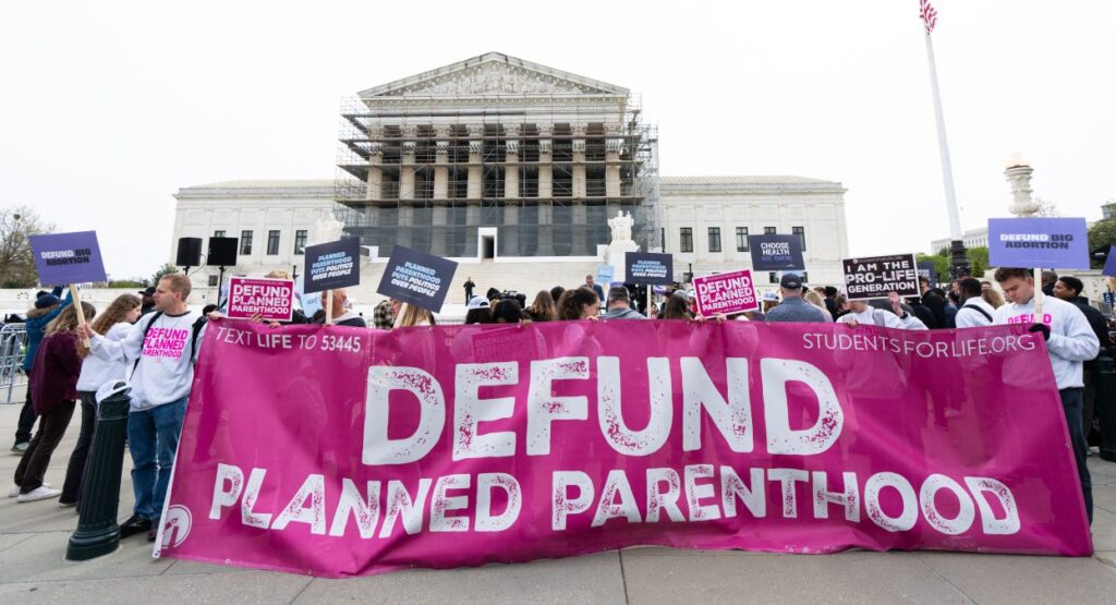 Pro-life supporters holding large "Defund Planned Parenthood" banner demonstrate in front of the Supreme Court.