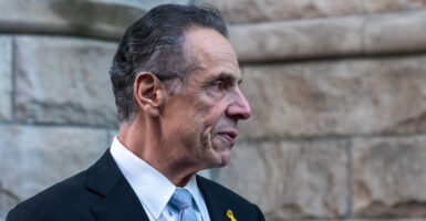 Andrew Cuomo stands wearing a black suit and light-blue necktie.