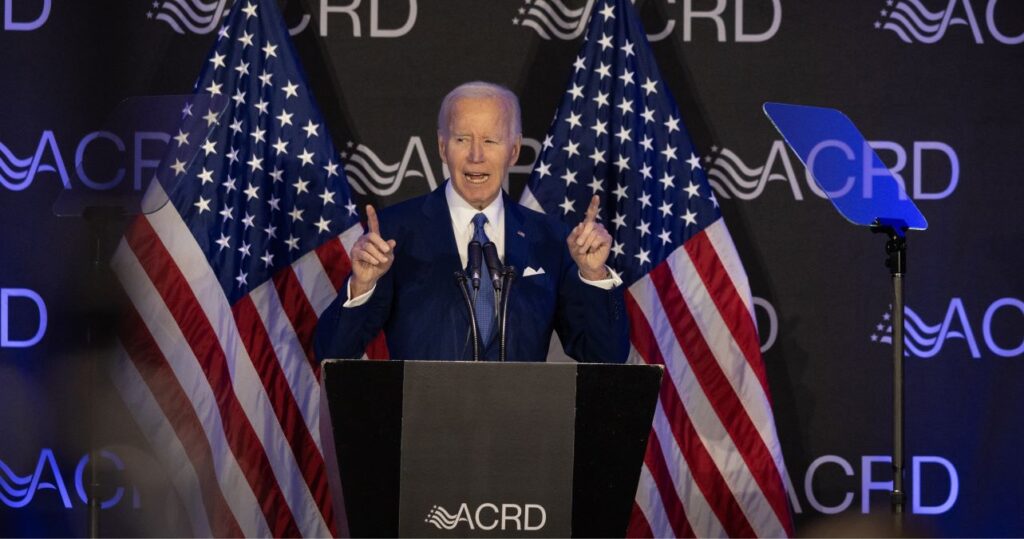 Joe Biden, fingers pointing upward, gives speech in front of two American flags.
