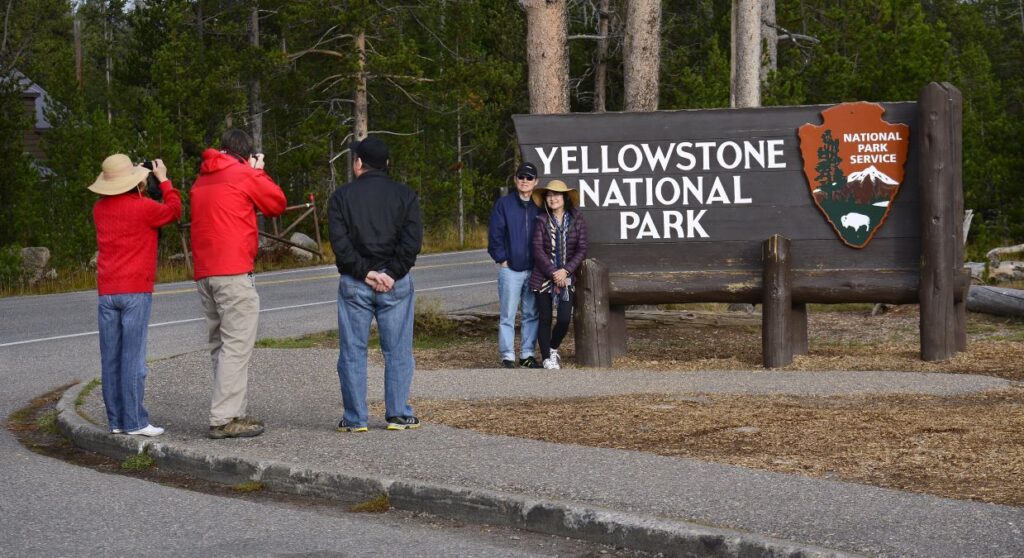 Two Chinese tourists pose in front of a sign for Yellowstone National Park, as two others snap pictures and one man, hands behind his back looks on.