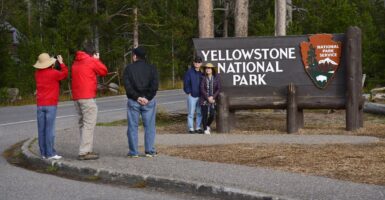 Two Chinese tourists pose in front of a sign for Yellowstone National Park, as two others snap pictures and one man, hands behind his back looks on.