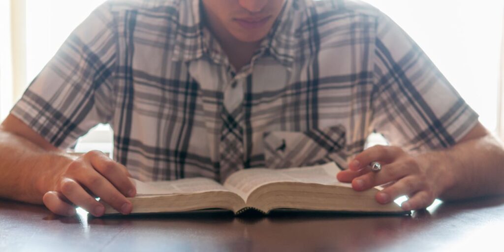 Teenage male sits at a desk reading, and studying the Bible, pen in hand.