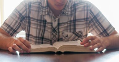 Teenage male sits at a desk reading, and studying the Bible, pen in hand.