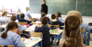 A shot from the back of a classroom of young students paying attention to their teacher. In the right foreground is a wooden sculpture of the Madonna and Child.