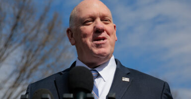 A smiling Tom Homan in a dark suit speaks into a microphone outdoors to reporters not seen in the photo