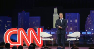 Jake Tapper standing in a black suit on a stage in front of a red CNN sign.