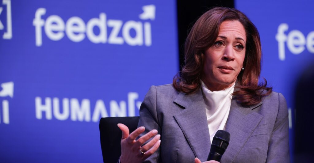 Kamala Harris sits in a chair with a microphone in her left hand and raises her right hand.