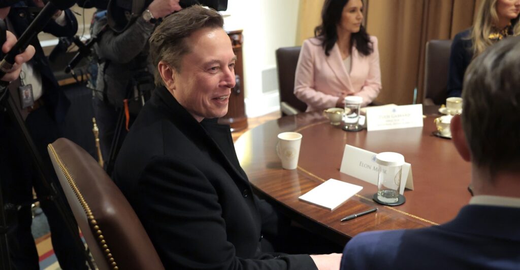 Elon Musk sits in a chair at a table and smiles as his head is turned to his right.