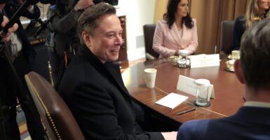 Elon Musk sits in a chair at a table and smiles as his head is turned to his right.