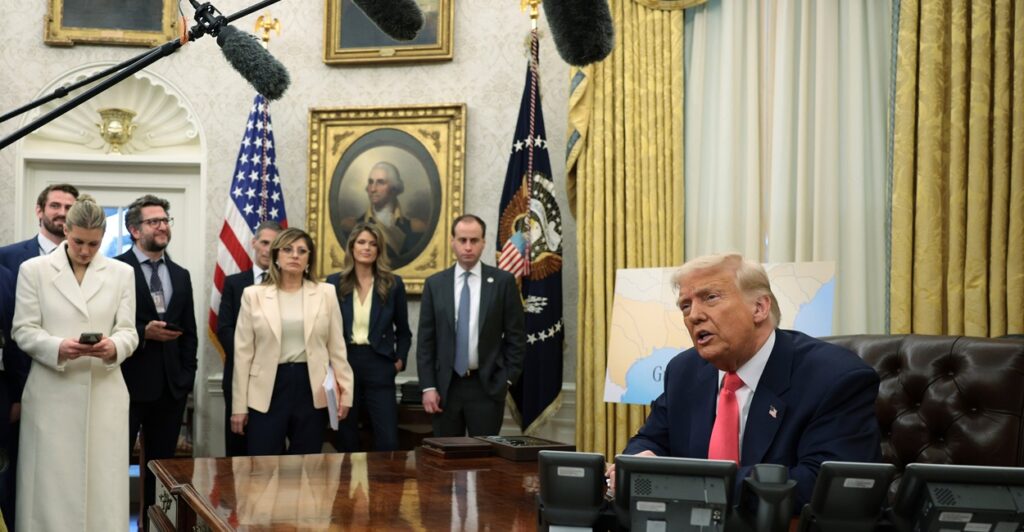 Donald Trump sits at his desk and talks to reporters stationed in front of him.