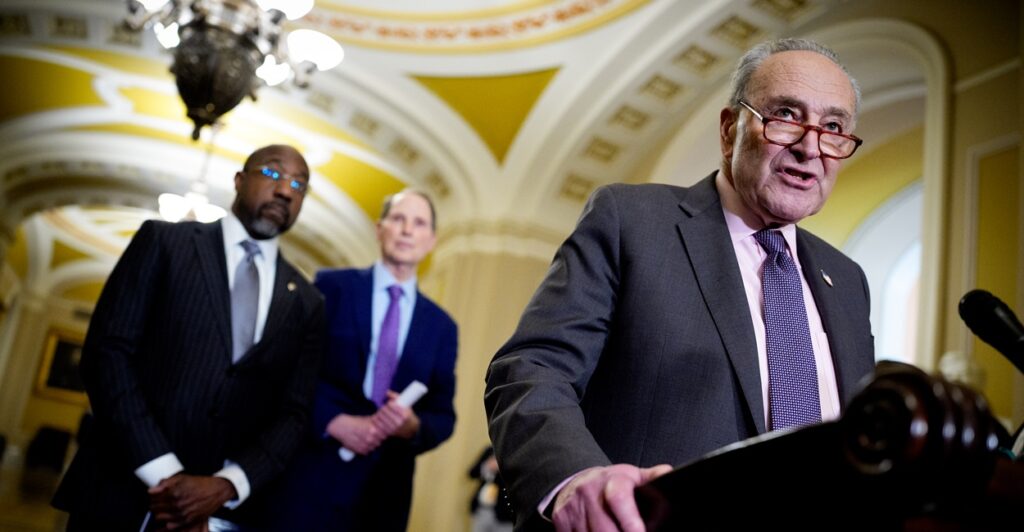 Chuck Schumer stands at a podium with his right hand gripping the podium as he prepares to speak to reporters.
