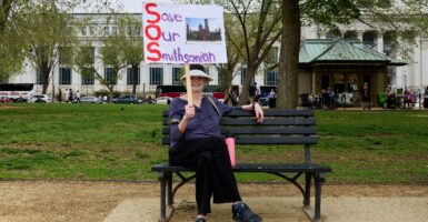 A woman sits on a park bench and holds a sign up in the air with her right hand. The sign states, "Save Our Smithsonian".