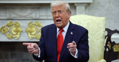 President Donald Trump sits in the Oval Office in a yellow chair wearing a dark blue suite and red tie. He is speaking and both his finders are pointed forward.