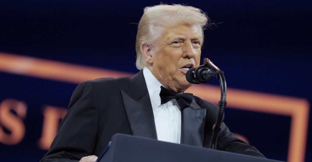 Donald Trump wears a tuxedo as he stands in front of a podium and speaks into a microphone.