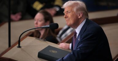 Donald Trump stands at a podium and looks to his left with a smirk on his face.