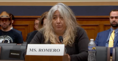 Cindy Romero, a woman with white hair and a black shirt, sits on a panel with a name tag in front of her.