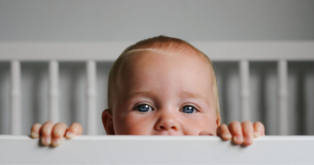 A baby in a crib, holding onto its rim to remain upright