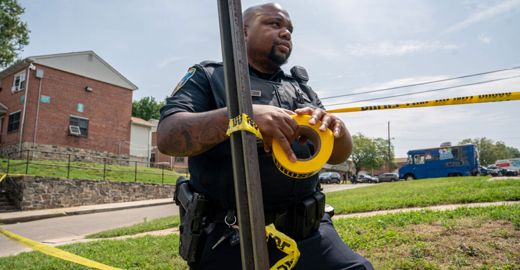 A black uniformed Baltimore Police officer hangs police tape at the site of a mass shooting