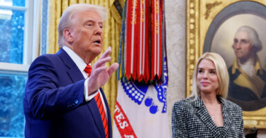 President Donald Trump in a blue suit and red tie gesturing with his hands while Attorney General Pam Bondi smiles and looks on. In the oval office.