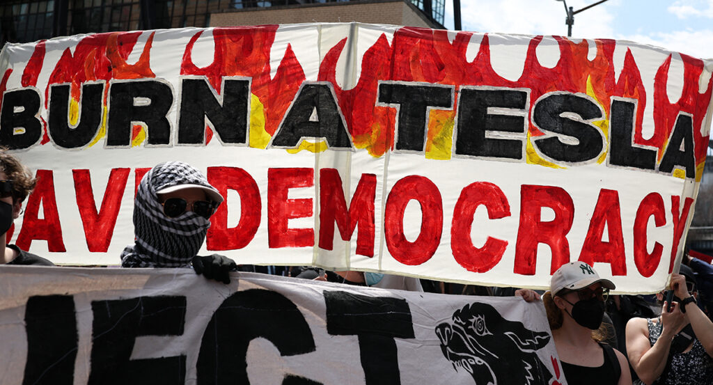 Protesters hold a sign reading, "Burn a Tesla, Save Democracy."