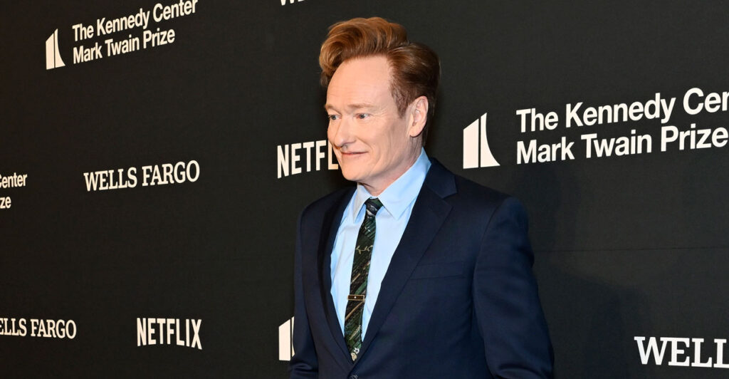 Conan O'Brien in a blue suit stands in a media photo op line