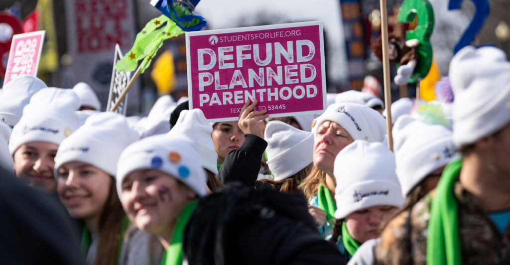 Protestors at the 2025 March for Life in white winter hats holding up Defund Planned Parenthood signs.