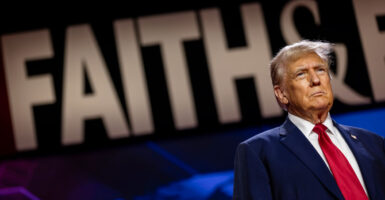 Donald Trump in a blue suit and red tie stands sternly in front of a large Faith sign in the background.