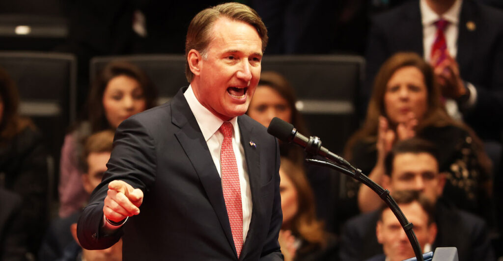 Virginia Governor Glenn Youngkin in a dark suit and orange tie speaking to an audience in gesturing by pointing his finger