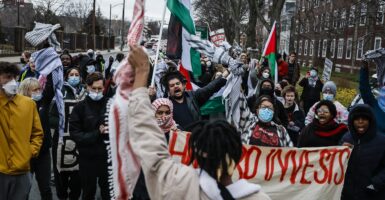 A group of students stand in the middle of the street and yell and wave flags as they protest.