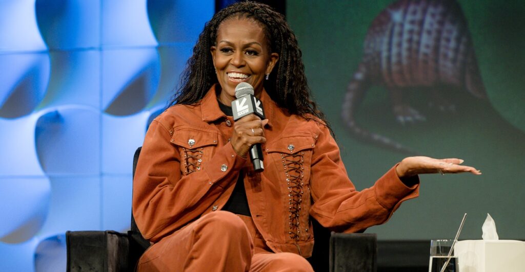 Michelle Obama sits in a chair and holds a microphone in her right hand as she holds out her left hand.