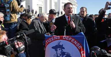 Nick Freitas in a suit and winter coat speaks forcefully into a microphone in the middle of a large crowd outside the Virginia Capitol. Draped on the front of the lectern is the Virginia Citizens Defense League (the gun rights group) banner