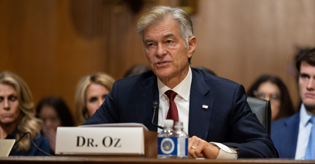 Dr. Mehmet Oz in a blue suit and red tie sitting at a witness table and microphone in a Senate hearing room