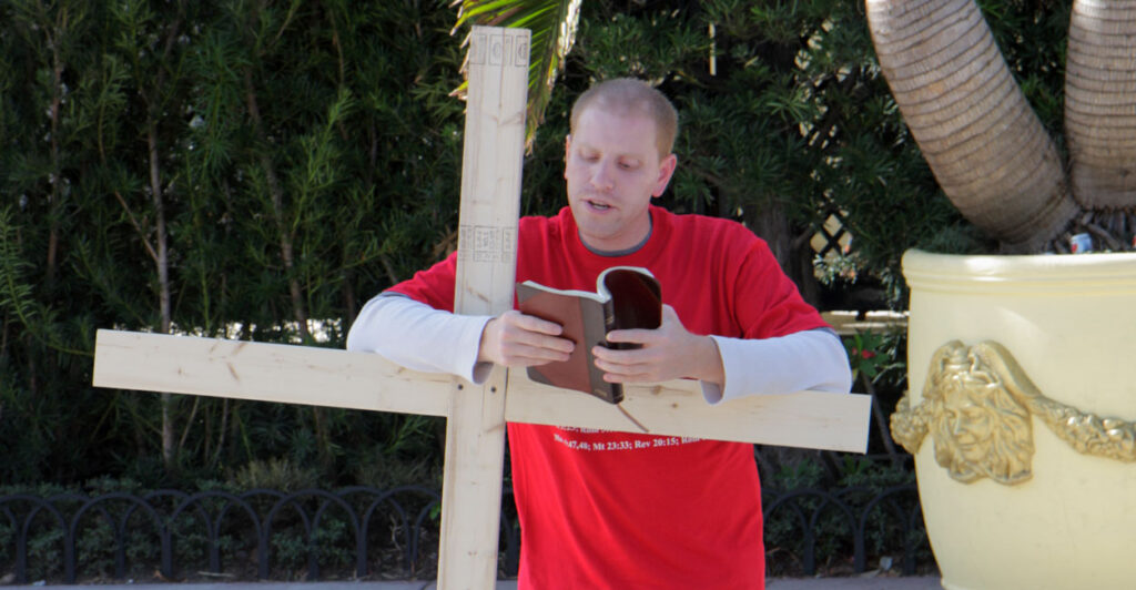 a young street preacher in a red T-shirt leaning on a wooden cross and reading from the Bible