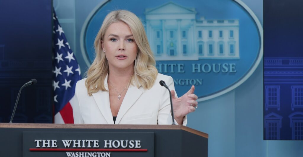 Karoline Leavitt stands behind a podium and holds up her left hand while talking to the press.