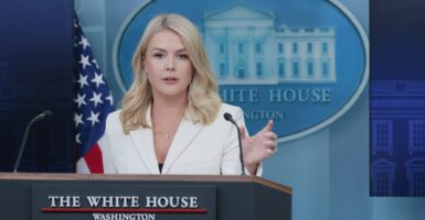 Karoline Leavitt stands behind a podium and holds up her left hand while talking to the press.