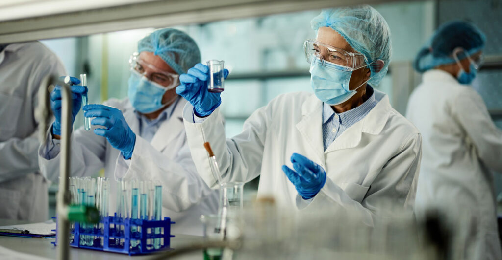 scientists in white robes and blue masks in a lab examining test tubes in beakers with liquids