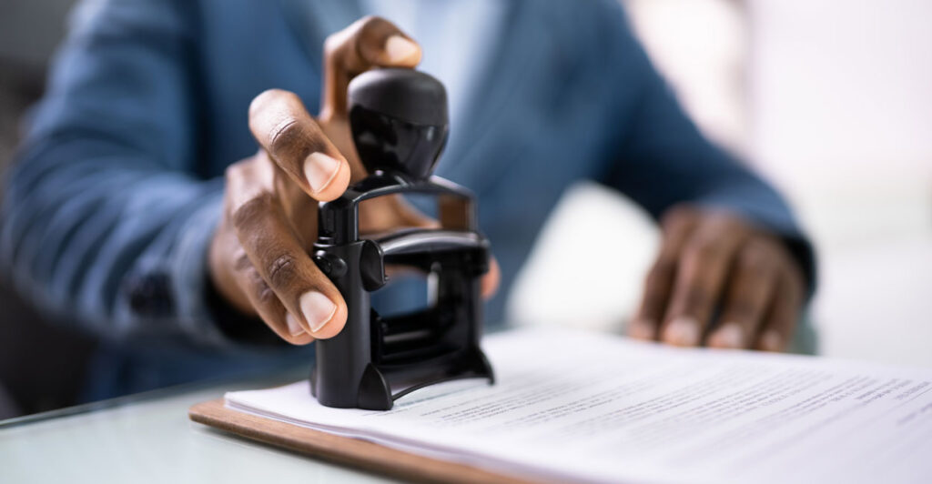 A black male Government worker in a blue sport jacket putting an official stamp on a government document