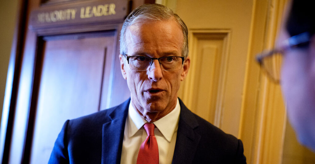 Senate Majority Leader John Thune in a blue suit and red tie in the hallway outside his office with his office door labeled majority leader in the background