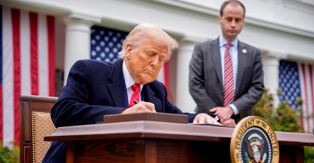 Donald Trump sits at a desk slightly bent over as he signs an executive order.