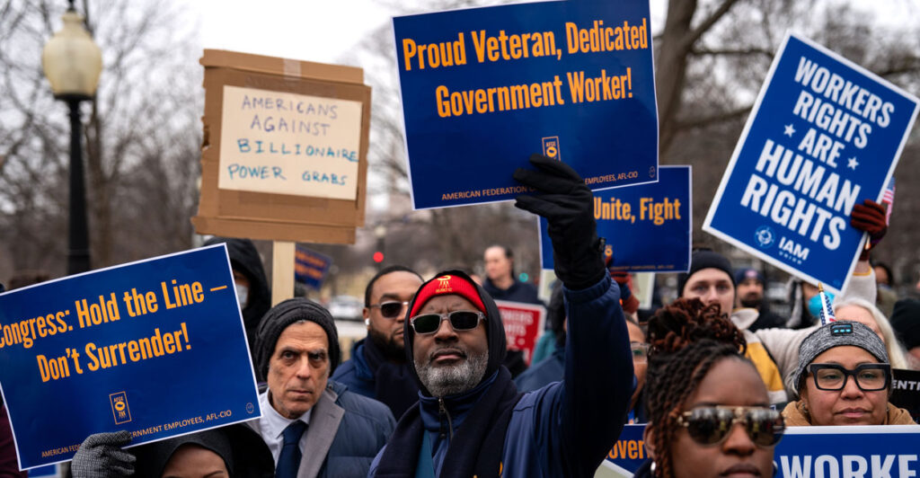 government workers protesting in the streets with signs such as "proud veteran, dedicated government worker" and "workers rights are human rights"