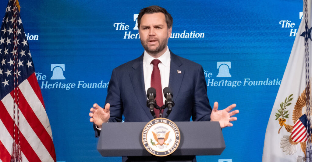 Vice President JD Vance in a blue suit and red tie at a lectern with microphones with an American flag in the background and the Heritage foundation logo in the background