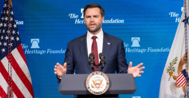 Vice President JD Vance in a blue suit and red tie at a lectern with microphones with an American flag in the background and the Heritage foundation logo in the background