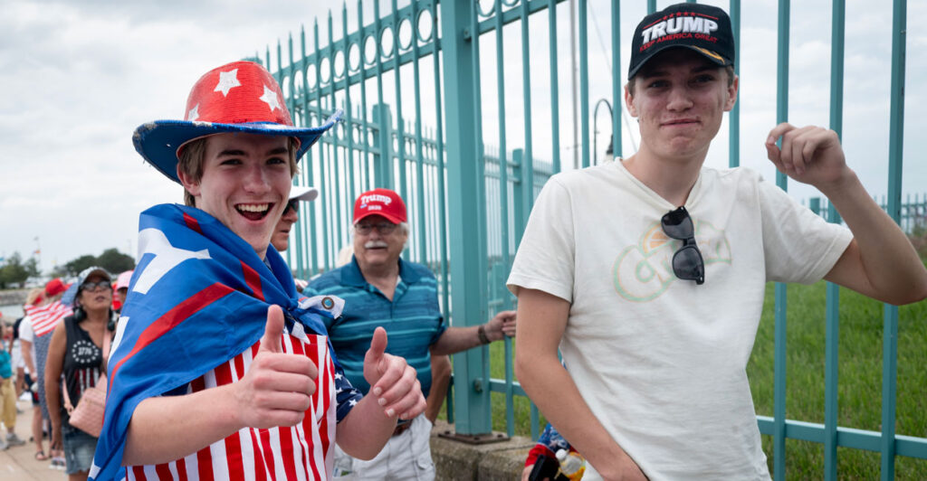 two young men wait to enter a Trump rally, one with a Trump hat on the other dressed in red white and blue with a Trump banner as a cape