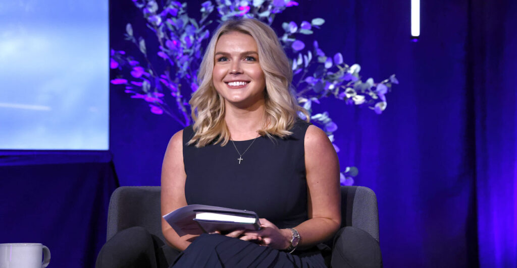 A smiling Karoline Leavitt in blue dress, in front of a backdrop of flowering tree.